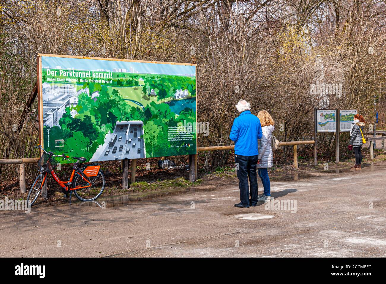 Eine Informationstafel im Englischen Garten in München, Deutschland, verkündet: "Der Parktunnel kommt." Zwei Besucher lesen die Details eines geplanten Straßentunnels, der den Verkehr unter dem Park hindurchführen soll, während ein rotes Mietfahrrad an der Tafel lehnt. Der Text dankt der Stadt München und dem Freistaat Bayern für das Projekt. Stockfoto