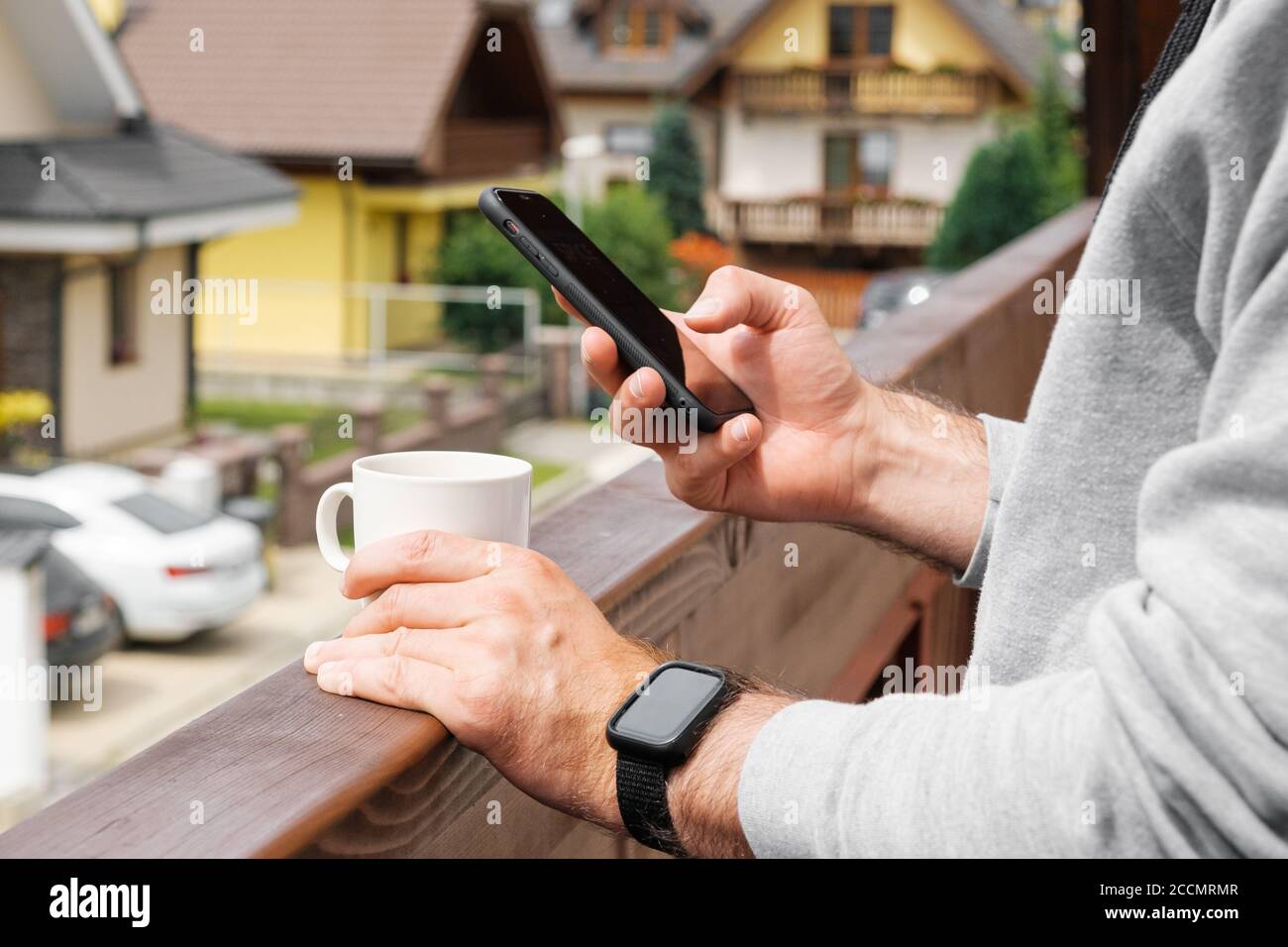 Mann in Smart Watch trinkt Kaffee oder Tee auf dem Balkon auf dem Hintergrund von Villen oder Pensionen, Nahaufnahme mans Hand mit seinem Smartphone. Konzept von Stockfoto Mann in Smart Watch trinkt Kaffee oder Tee auf dem Balkon auf dem Hintergrund von Villen oder Pensionen, Nahaufnahme mans Hand mit seinem Smartphone. Konzept von Stockfoto