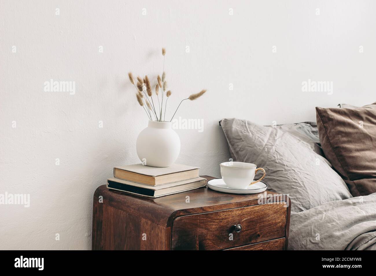 Moderne weiße Keramikvase mit trockenem Lagurus ovatus Gras und Tasse Kaffee auf Retro-Nachttisch aus Holz. Beigefarbene Bettwäsche und Samtkissen im Schlafzimmer Stockfoto