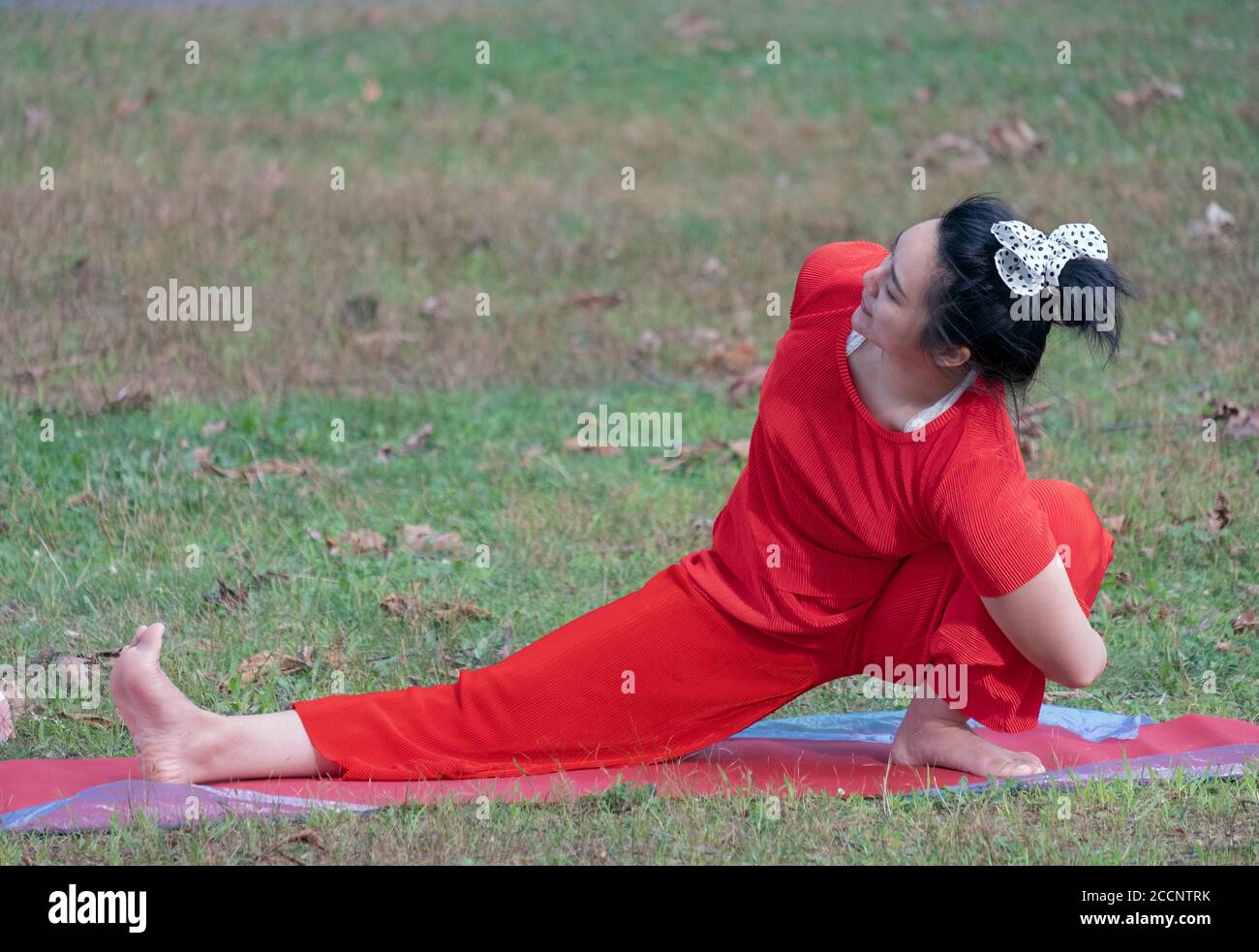 Eine Frau, die einen roten Hosenanzug und einen Haarbogen mit Polka-Punkt trägt, macht einen schwierigen Stretcn bei einem Yoga-Kurs in einem Park in Queens, New York Stockfoto