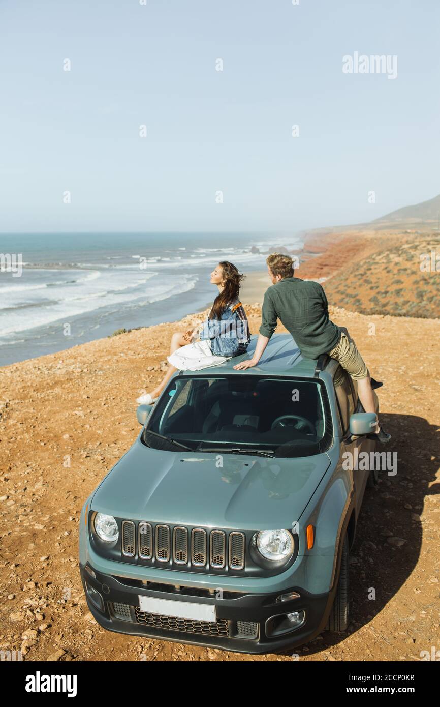 Roadtrip-Konzept. Junges und glückliches Hippie-Paar, das auf dem Dachauto sitzt und einen fantastischen Blick auf das Meer und die Küste Marokkos genießt. Fernweh und Lifestyle. Stockfoto