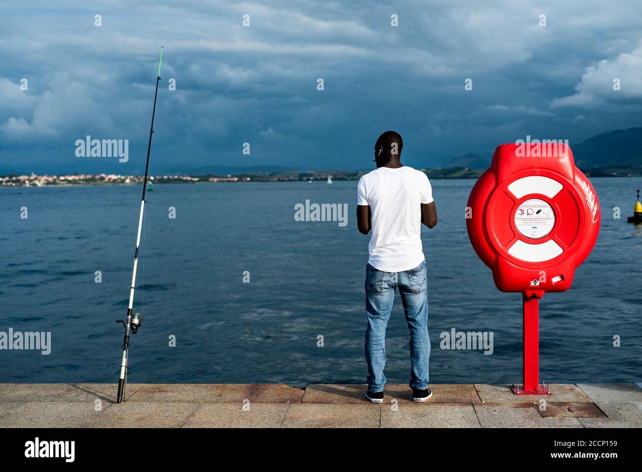 Rückansicht eines jungen schwarzen Mannes in einem weißen T-Shirt. Schwarzer Mann, der beim Angeln in Paseo Maritimo, Santander, Spanien Musik hört Stockfoto