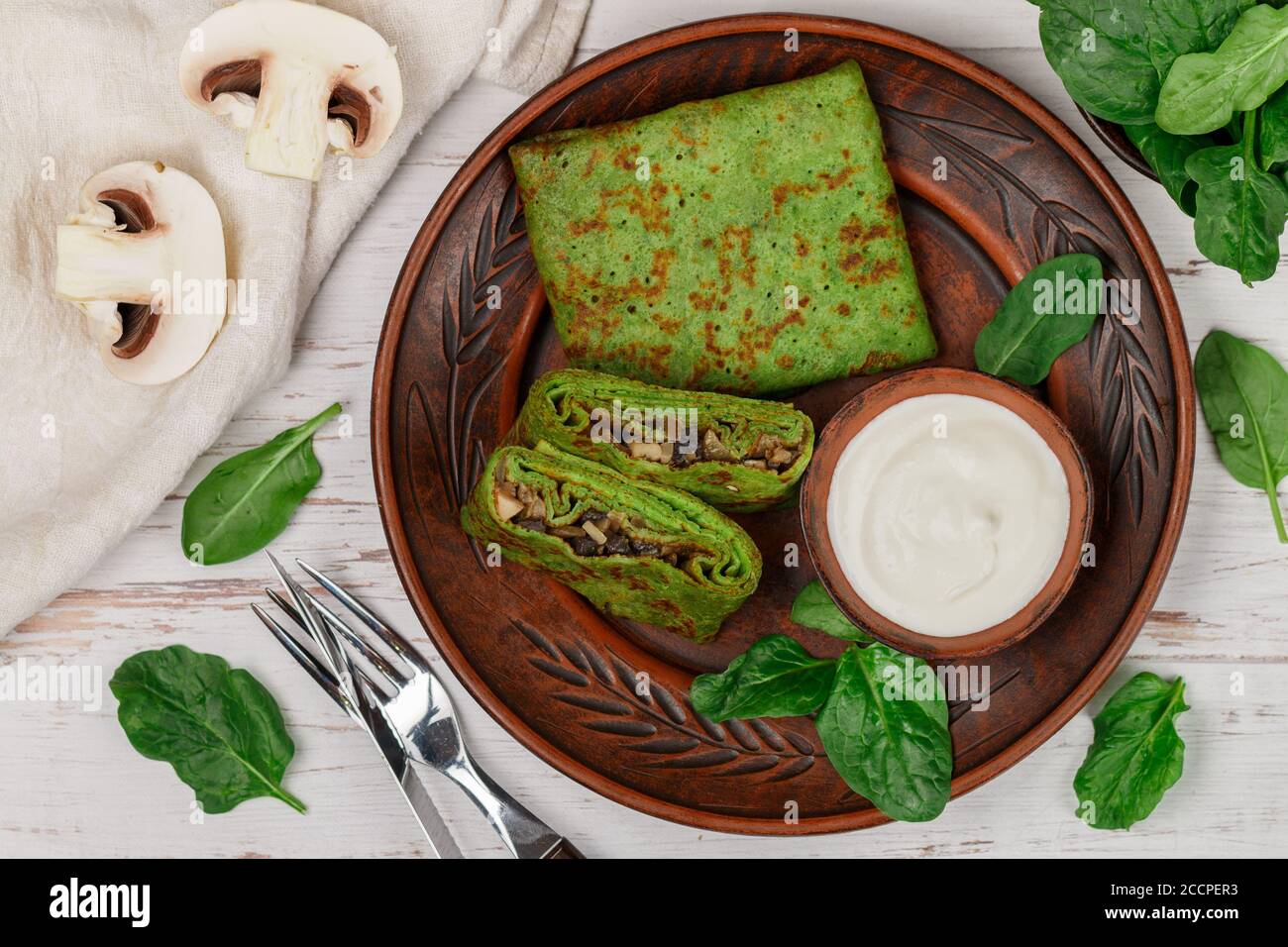Spinat grüne Pfannkuchen (Crepes) gefüllt mit Pilzfüllung mit saurer Sahne. Köstliches gesundes Frühstück in einem Tonteller auf einem hellen Holzbackgroun Stockfoto