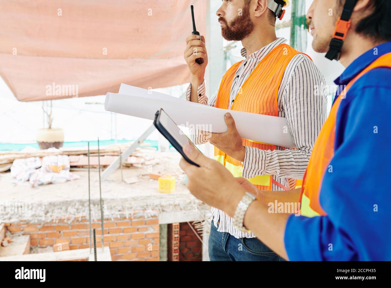 Bauingenieur mit Blaupause im Gespräch auf Walkie-Talkie, wenn sein Kollege In der Nähe stehend mit digitalem Tablet in der Hand Stockfoto