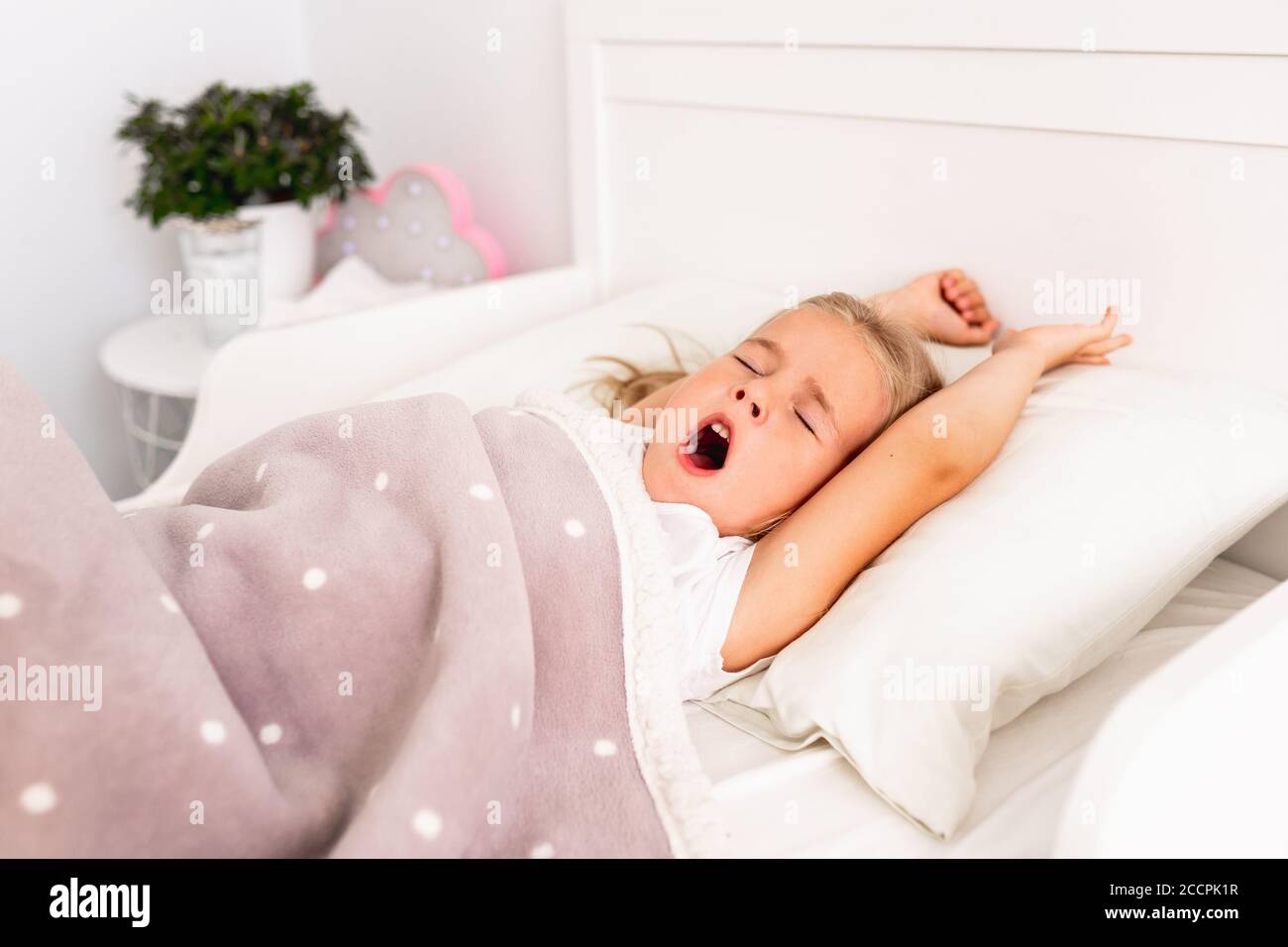 Draufsicht auf kleine niedliche blonde Mädchen schlafen auf weißem Bett mit ihren Händen nach oben. Kleinkind gähnt und will nicht aufwachen, um zur Schule zu gehen. Stockfoto