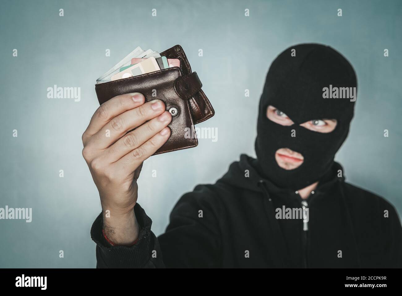 Der junge, unwillige Mann mit schwarzer Maske hält die schwarze Leder-Männer-Geldbörse und schaut zu seiner Beute. Räuber hat die Brieftasche gestohlen. Halb Schuss Dieb in Stockfoto