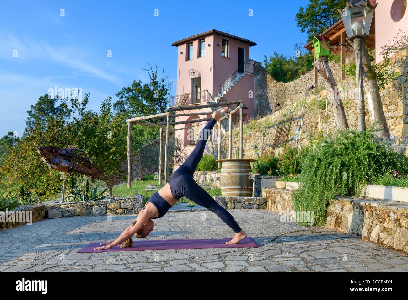 Mädchen tun einen nach unten Hund einzelnes Bein heben Yoga Pose Während eines morgendlichen Trainings auf einer gepflasterten Steinterrasse im Freien Im Garten in einem Gesundheits-und Fitness-Co Stockfoto
