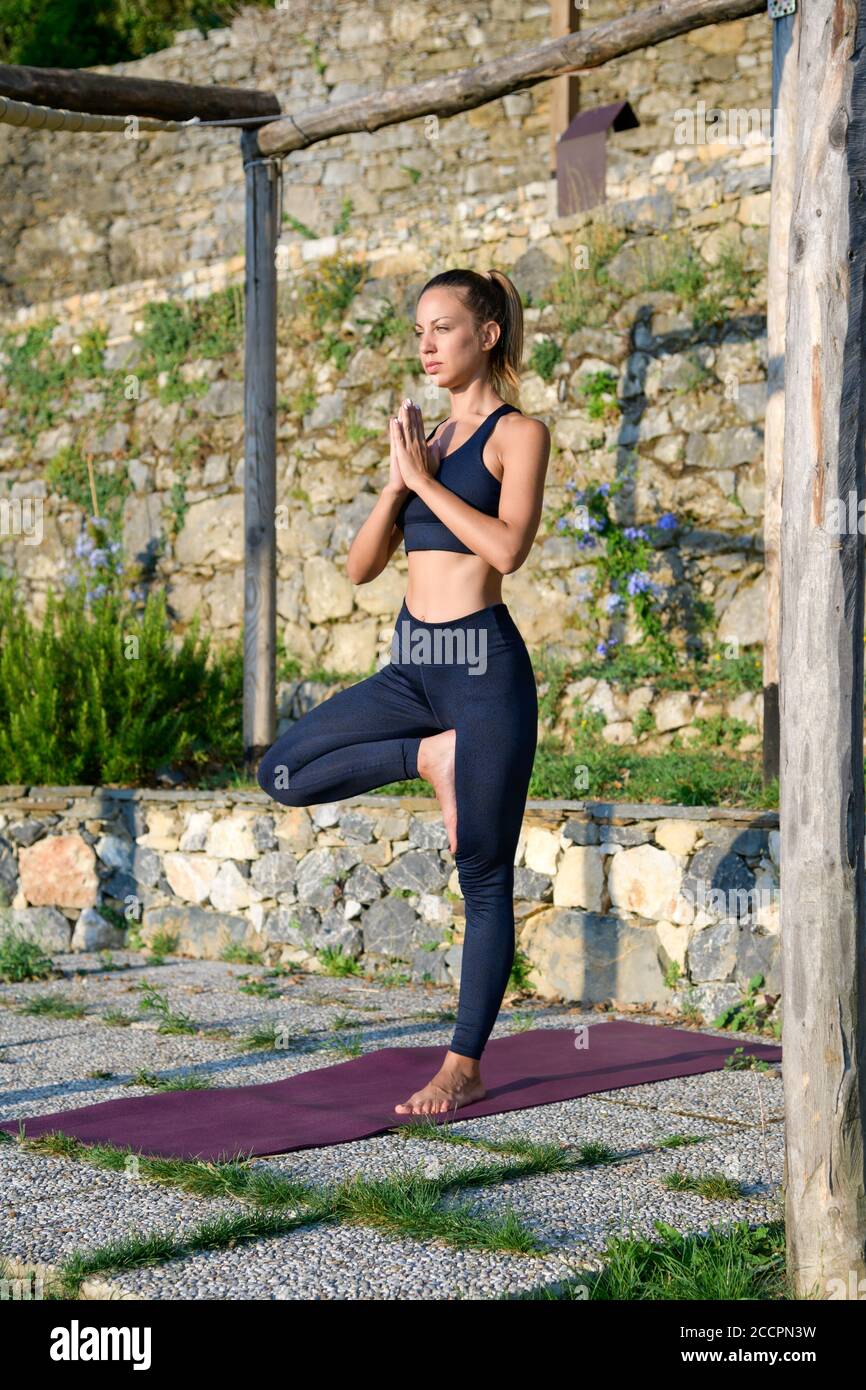 Frau, die eine Yoga-Baum-Pose ausarbeitet, ist ausgeglichen Ein Bein in der Morgensonne draußen im Garten innen Ein Gesundheits- und Fitnesskonzept Stockfoto