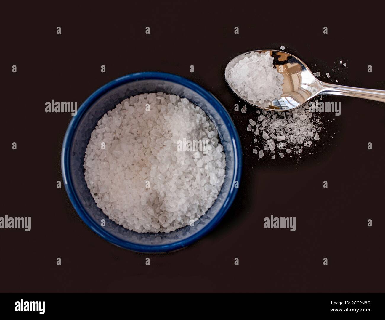 Salz in einem blauen Glas und einem silbernen Löffel mit weißen Salzkristallen. Flache Ansicht oben von oben auf schwarzem Hintergrund. Stockfoto