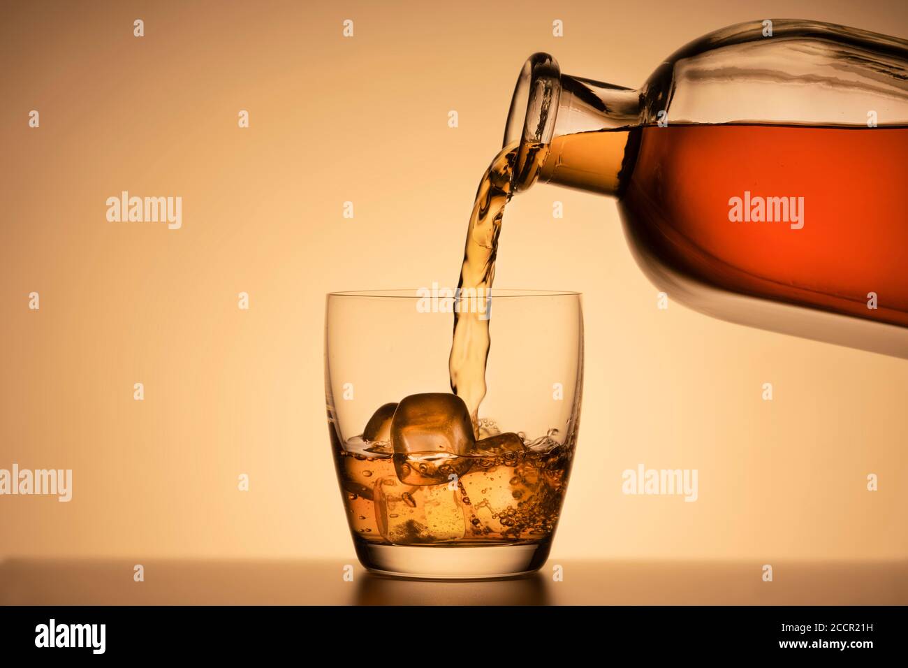 Ein Glas Whiskey auf den Felsen. Ausgiessen von Bourbon oder Cognac aus einer Spirituosenflasche. Oranger Hintergrund Stockfoto
