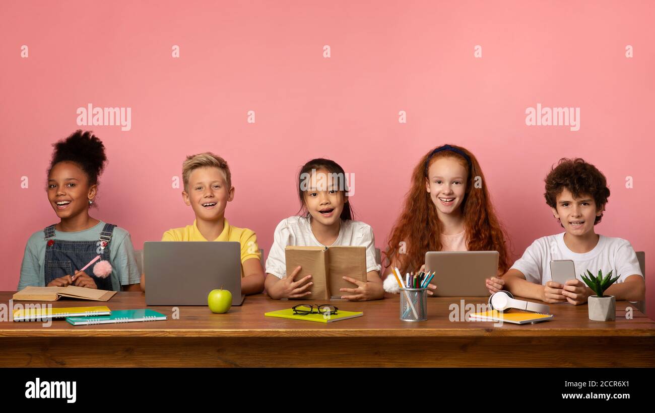 Gruppe von multiethnischen Schülern mit Büchern und elektronischen Geräten sitzen am Tisch über rosa Hintergrund, Leerraum Stockfoto