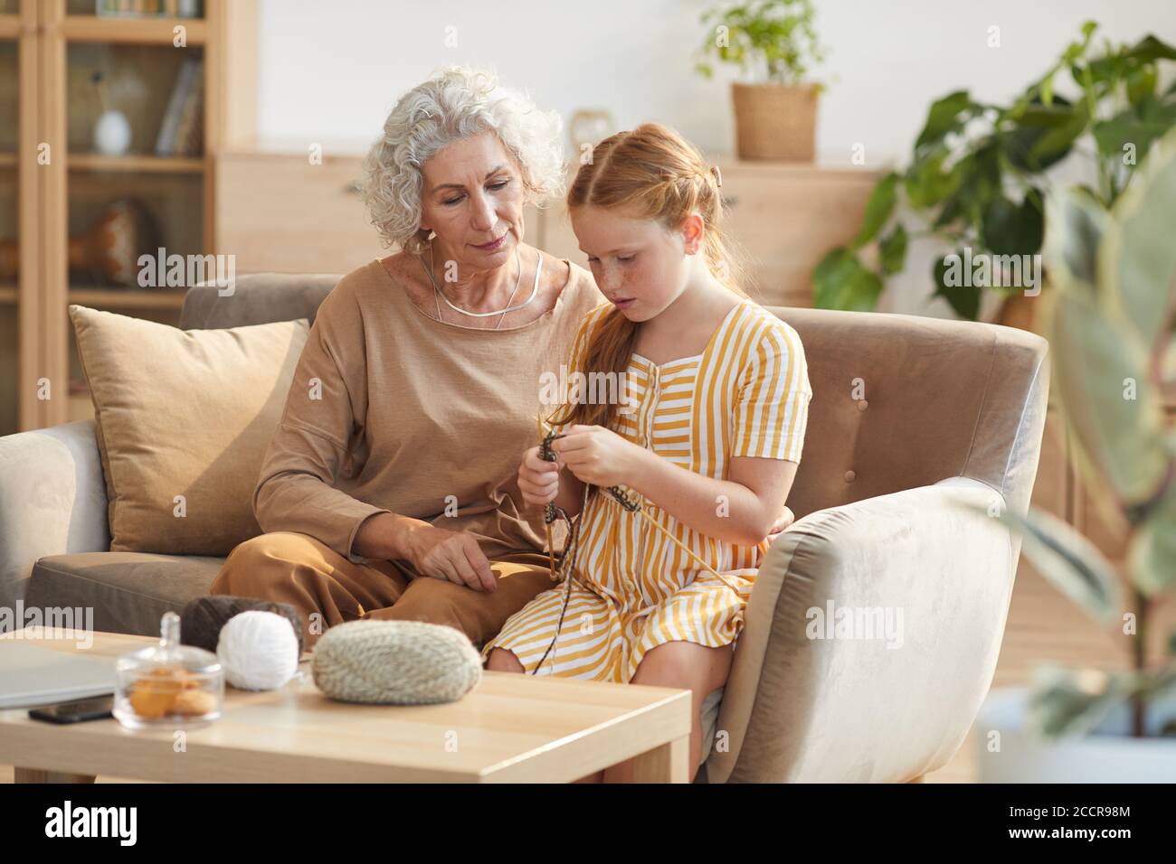 Warm getönte Porträt von eleganten Senior Frau Lehre Enkelin Stricken, während auf der Couch sitzen in gemütlichen Hause durch Sonnenlicht beleuchtet, kopieren Raum Stockfoto