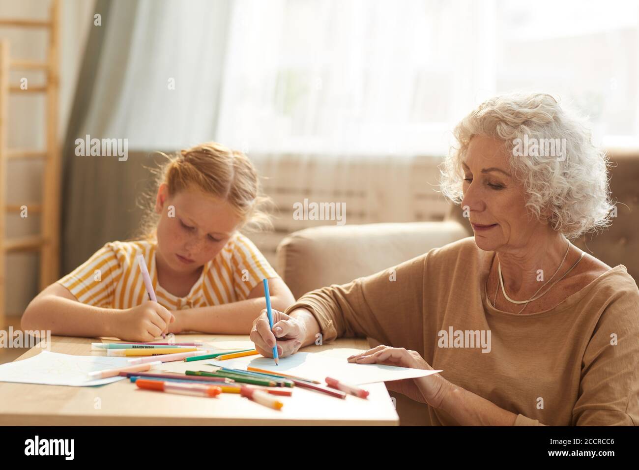 Warm getönte Porträt von lächelnden Senior Frau Babysitting niedlich rot Behaarte Mädchen und Zeichnung zusammen, während sitzen von Couchtisch In gemütlichem Wohnzimmer Stockfoto