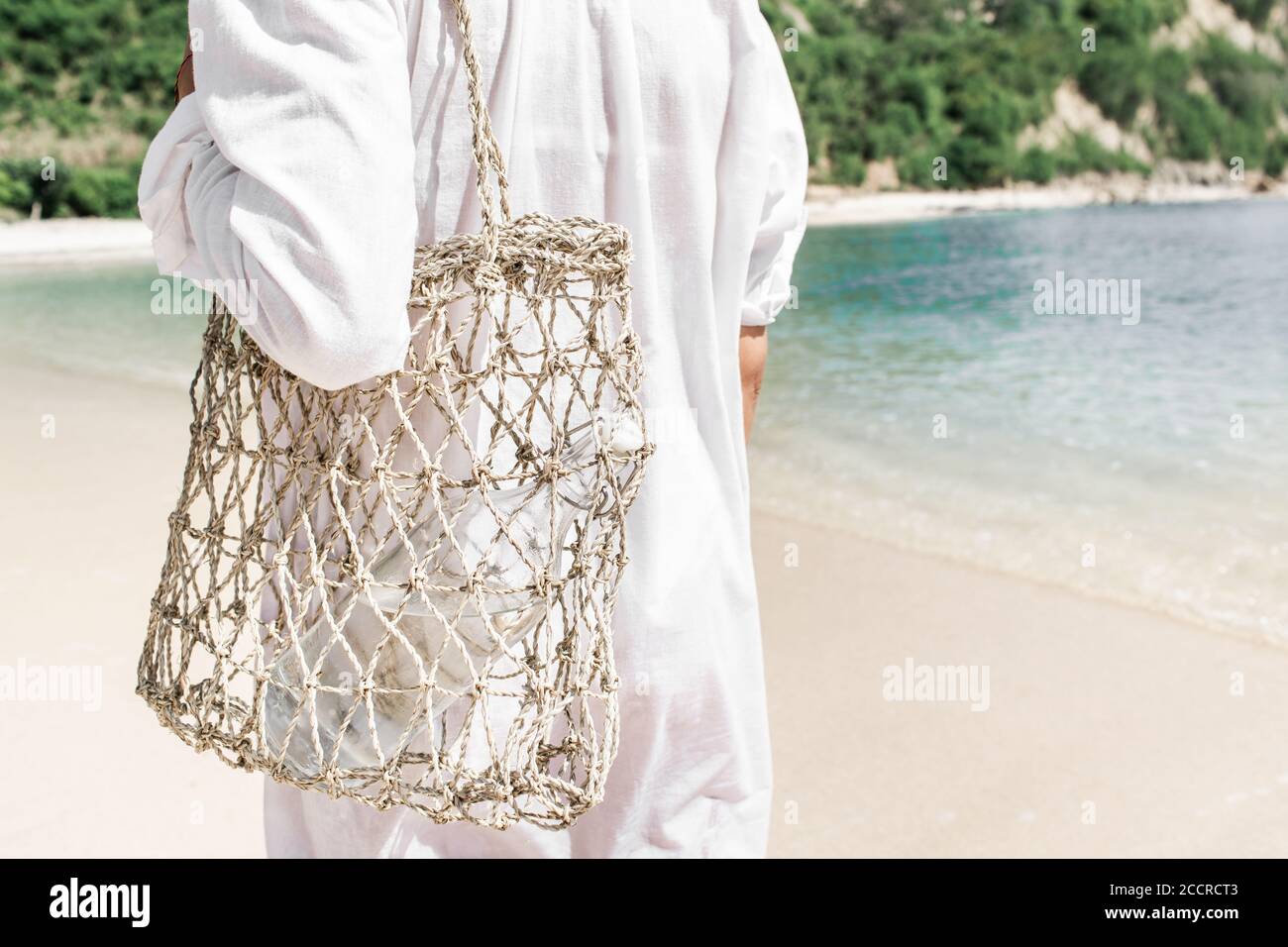 Frau hält Stroh Weidenwiederverwendbare Tasche mit Glasflasche für Wasser. Plastik frei und Zero Waste Lifestyle. Weißer Sandstrand mit klarem türkisfarbenem Wasser im Hintergrund. Stockfoto