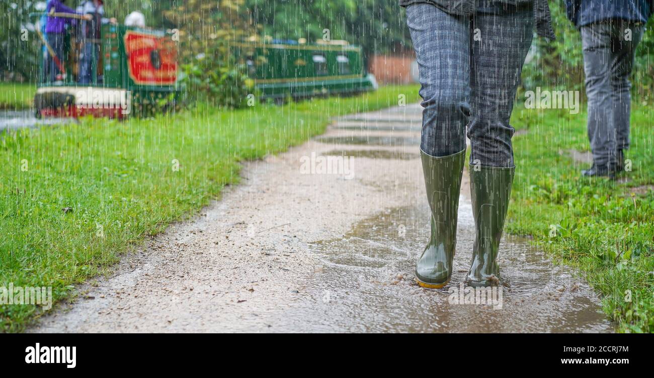 Nahaufnahme, Vorderansicht der grünen Gummistiefel, die von einer Frau getragen werden, die bei starkem Regen durch Pfützen spritzt, Großbritannien, auf einem Kanalpfad entlang. Stockfoto
