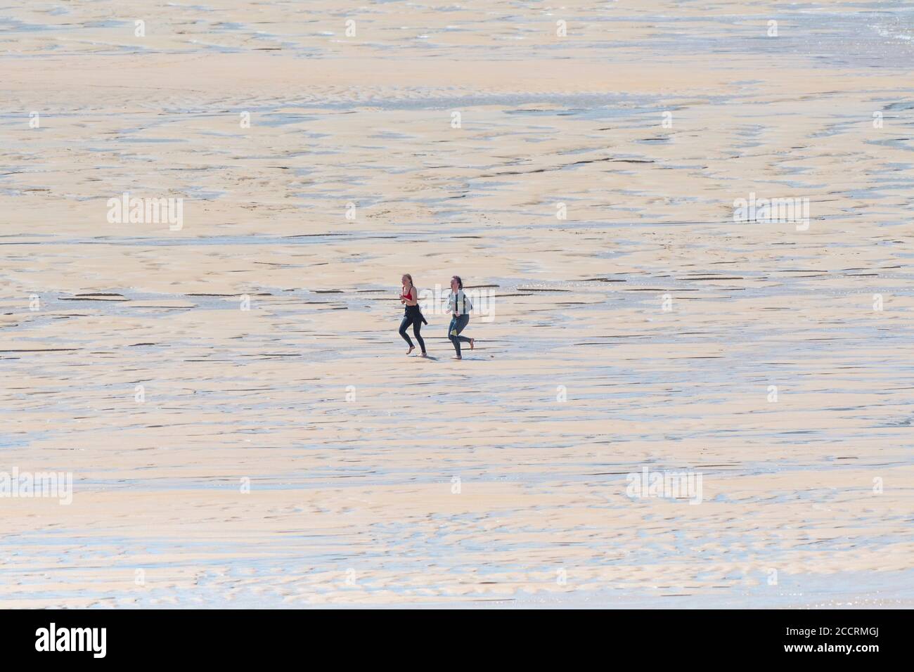 Ein paar von zwei jungen Weibchen in Neoprenanzügen, die über den Fistral Beach in Newquay in Cornwall laufen. Stockfoto