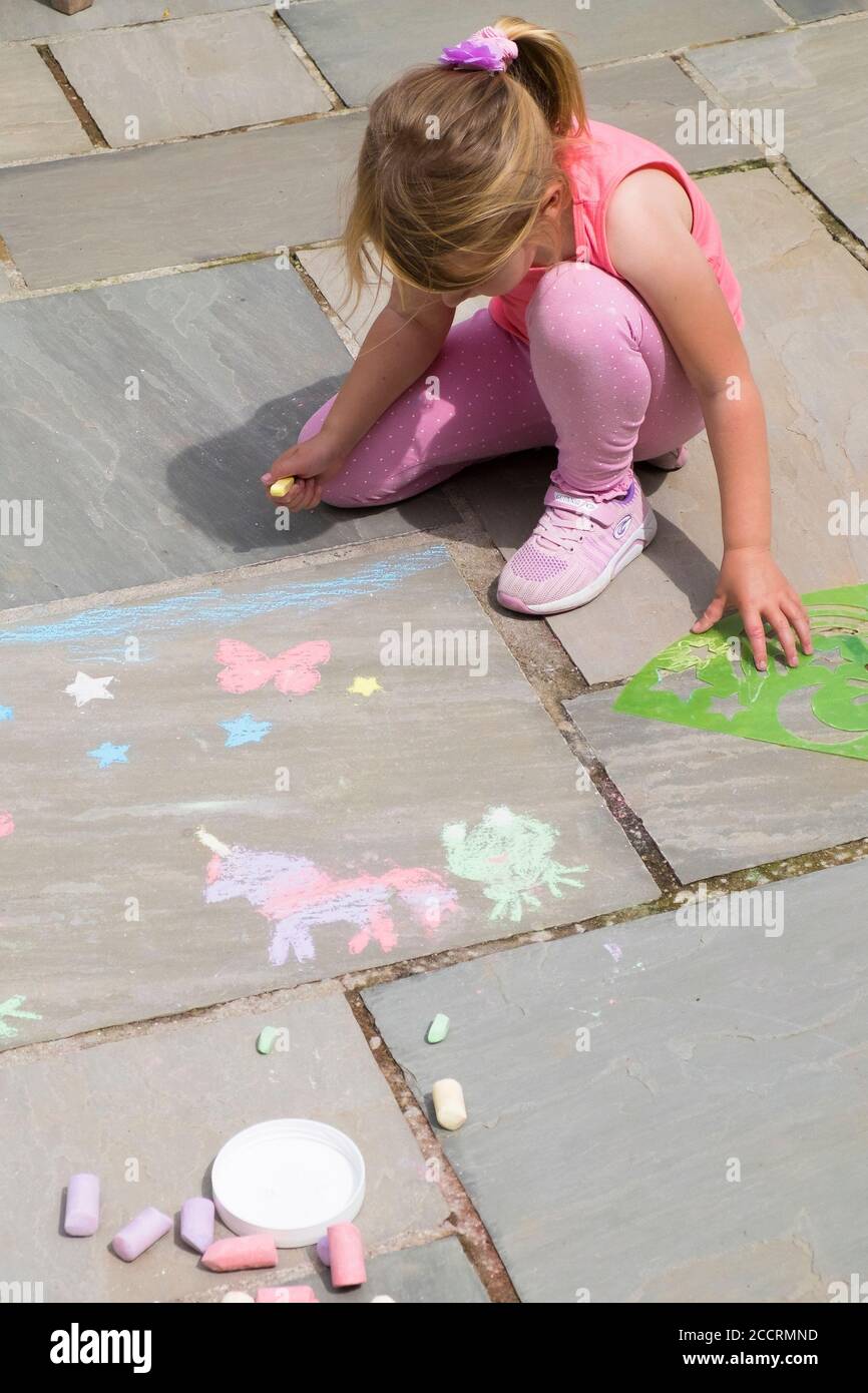 Ein fünfjähriges Mädchen amüsant herslef eifrig mit Plastikschablonen zu Kreide Formen auf einem Plattenboden. Stockfoto