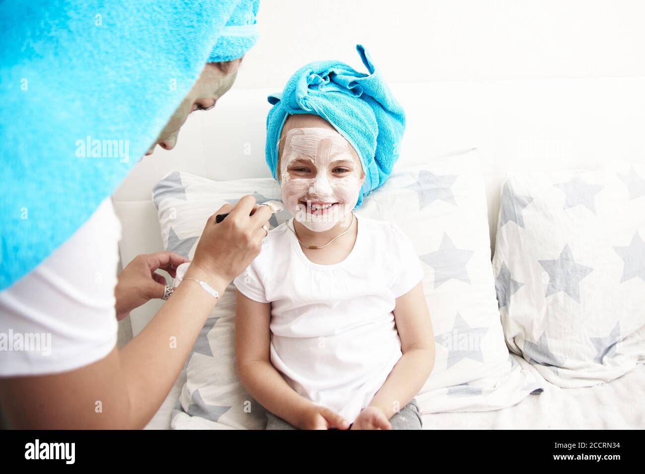 Nette junge Mutter und ihre Tochter sitzen auf dem Bett in Handtüchern auf ihrem Kopf und machen eine Gesichtsmaske. Hautpflege, Familienbeziehungen. Stockfoto