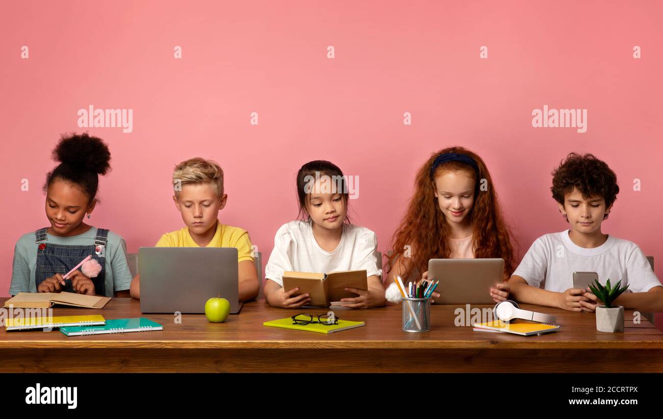 Multiethnische Kinder mit elektronischen Geräten und Büchern studieren am Tisch über rosa Hintergrund, freien Raum Stockfoto