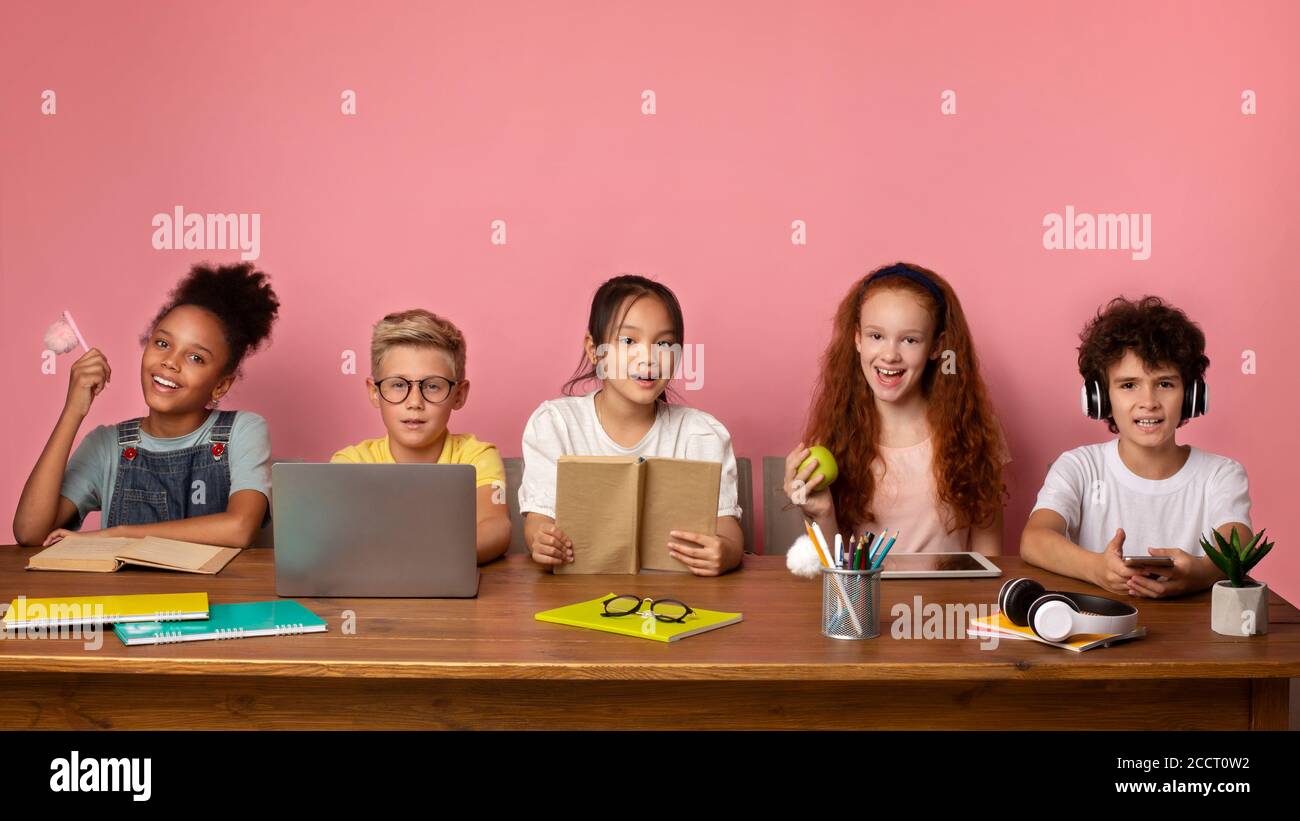 Bereit für die Schule. Diverse Jungen und Mädchen mit Lernmaterialien und Gadgets sitzen am Schreibtisch, rosa Hintergrund. Freier Speicherplatz Stockfoto