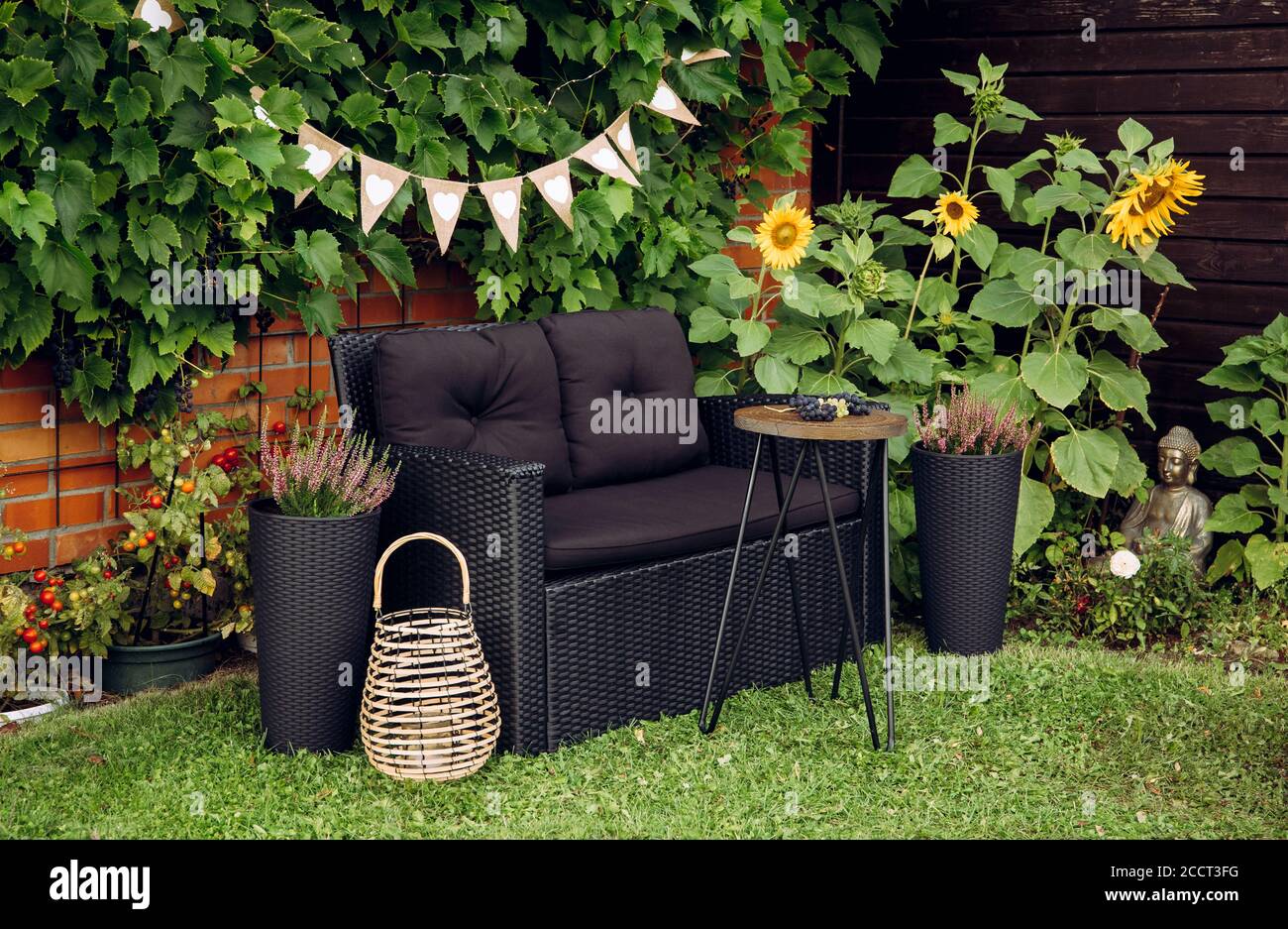 Schwarzer moderner Poly Rattan Gartenmöbelstuhl, Beistelltisch aus Metalldraht. Grüne Weinreben Pflanzen wachsen auf roten Backsteinmauer als dekoratives Element. Holz Stockfoto