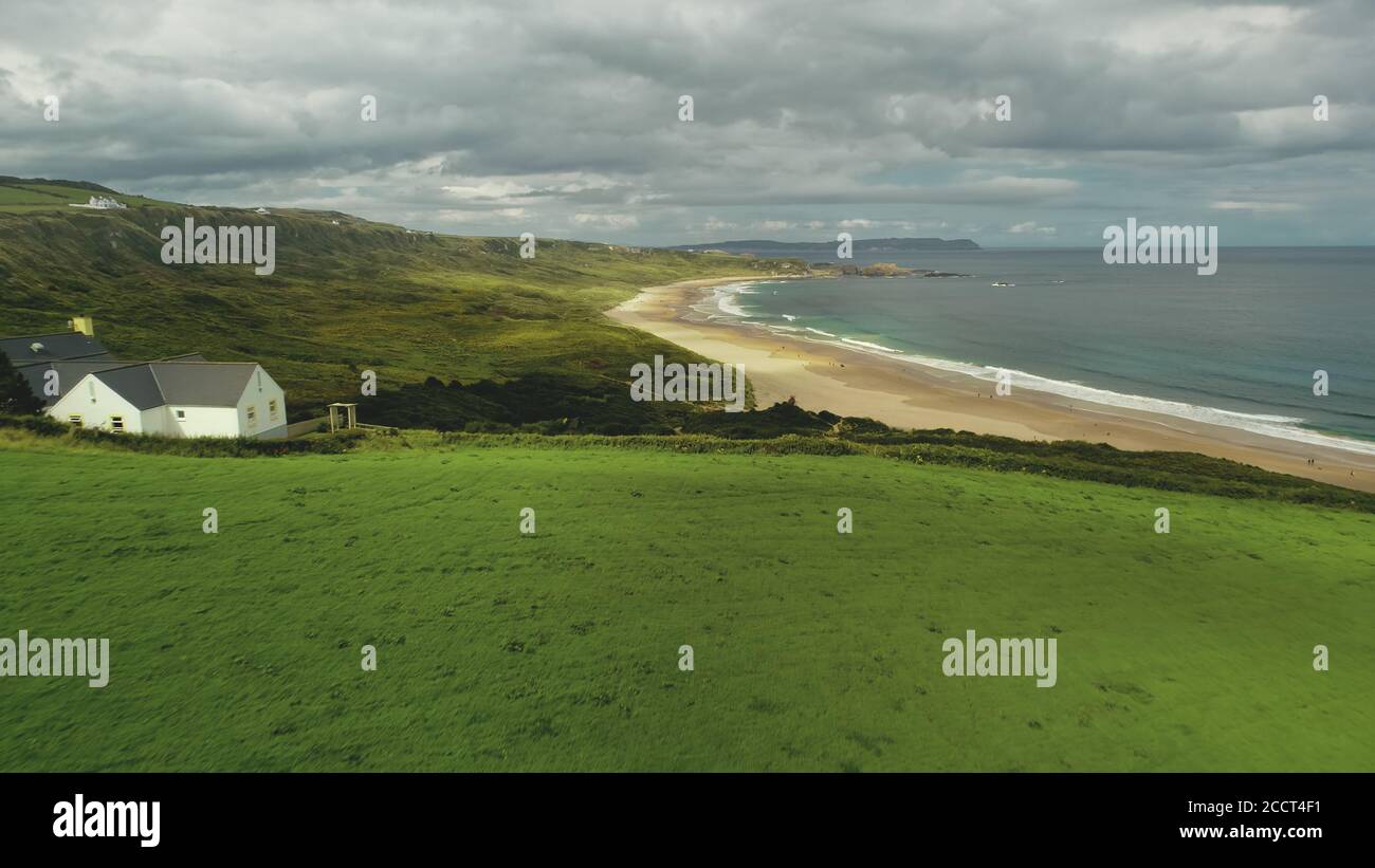 Luftaufnahme der Strandhütte: Grüne Wiese in der Nähe der Meeresbucht und einsame weiße Felsen. Graue Wolken schweben über Ozeanwasser, Sandküste. Ruhige Naturlandschaft in Nordirland. Filmmaterial Stockfoto