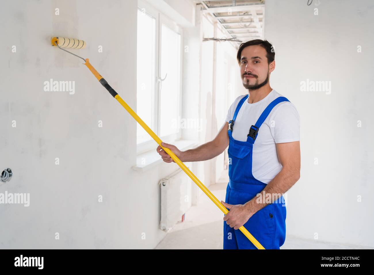 Ein bärtiger Mann in einem blauen Overall malt eine Wand im Flur. Er benutzt eine Walze Stockfoto