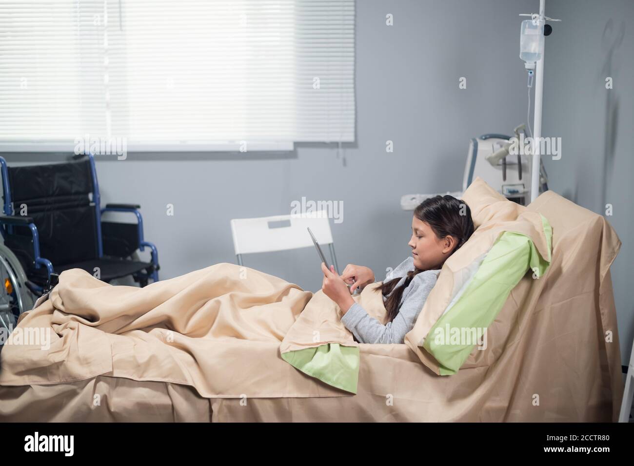 Kleines Mädchen mit einem Eis und einer Tablette in einem Krankenhausbett, ihr Rollstuhl ist neben ihrem Bett. Stockfoto