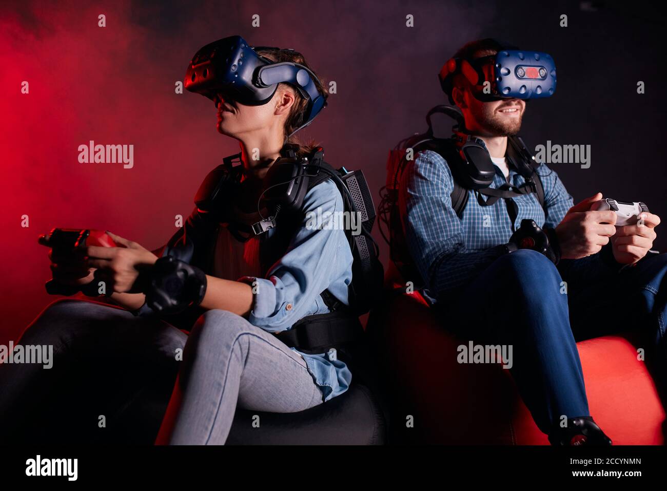 Kaukasische Männer und Frauen sitzen in Headsets, Brillen, Videospiele spielen, Joysticks halten. Rote Neon-Hintergrundbeleuchtung Stockfoto