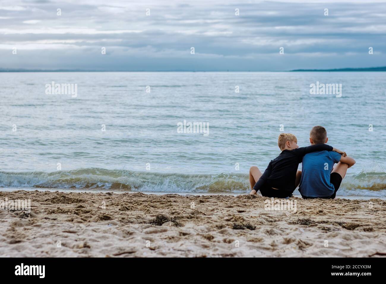 Zwei Brüder verbinden sich über die Schönheit des Ozeans, während sie am Strand sitzen, und der jüngere Bruder tröstet den älteren mit einem Arm um ihn herum. Stockfoto