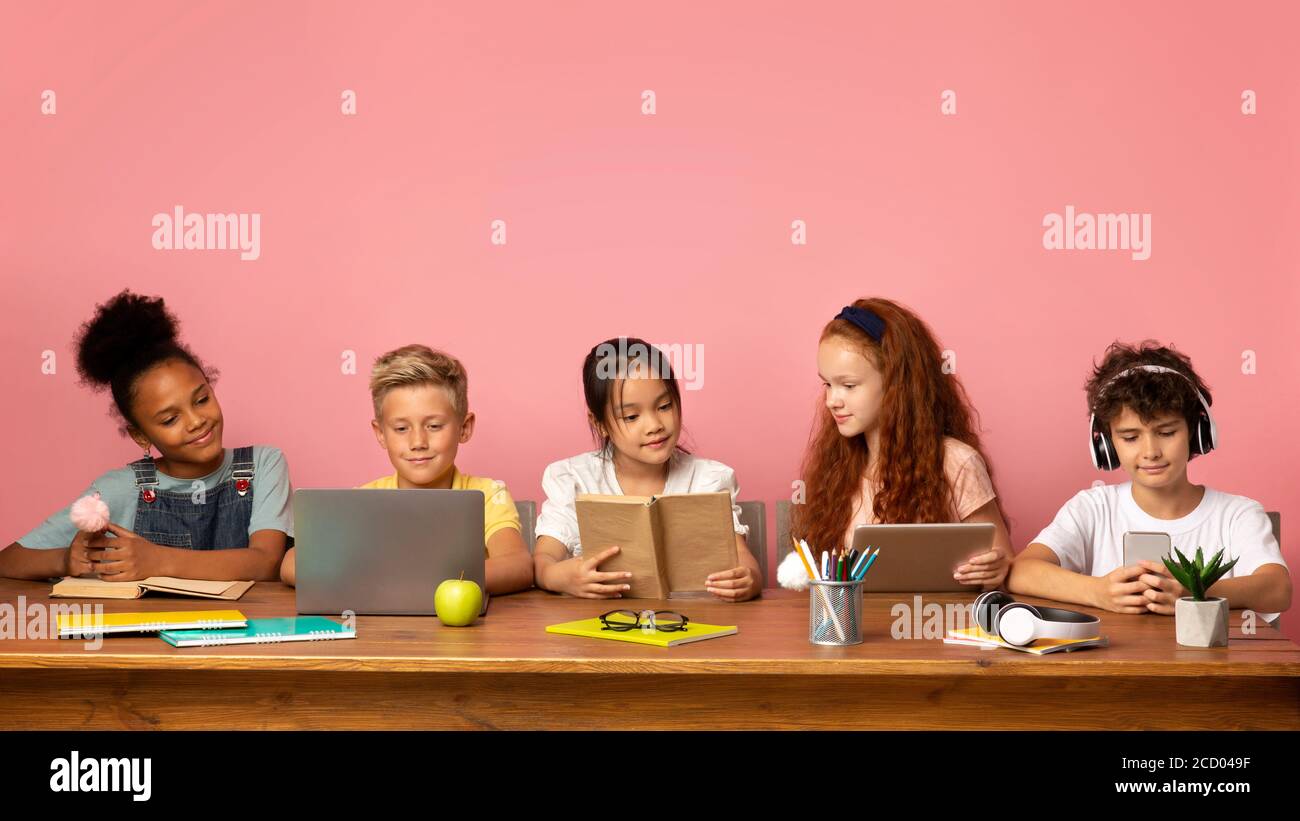 Schulzeit. Multiethnische Kinder mit verschiedenen Gadgets und Bücher sitzen am Tisch vor rosa Hintergrund, leerer Raum Stockfoto