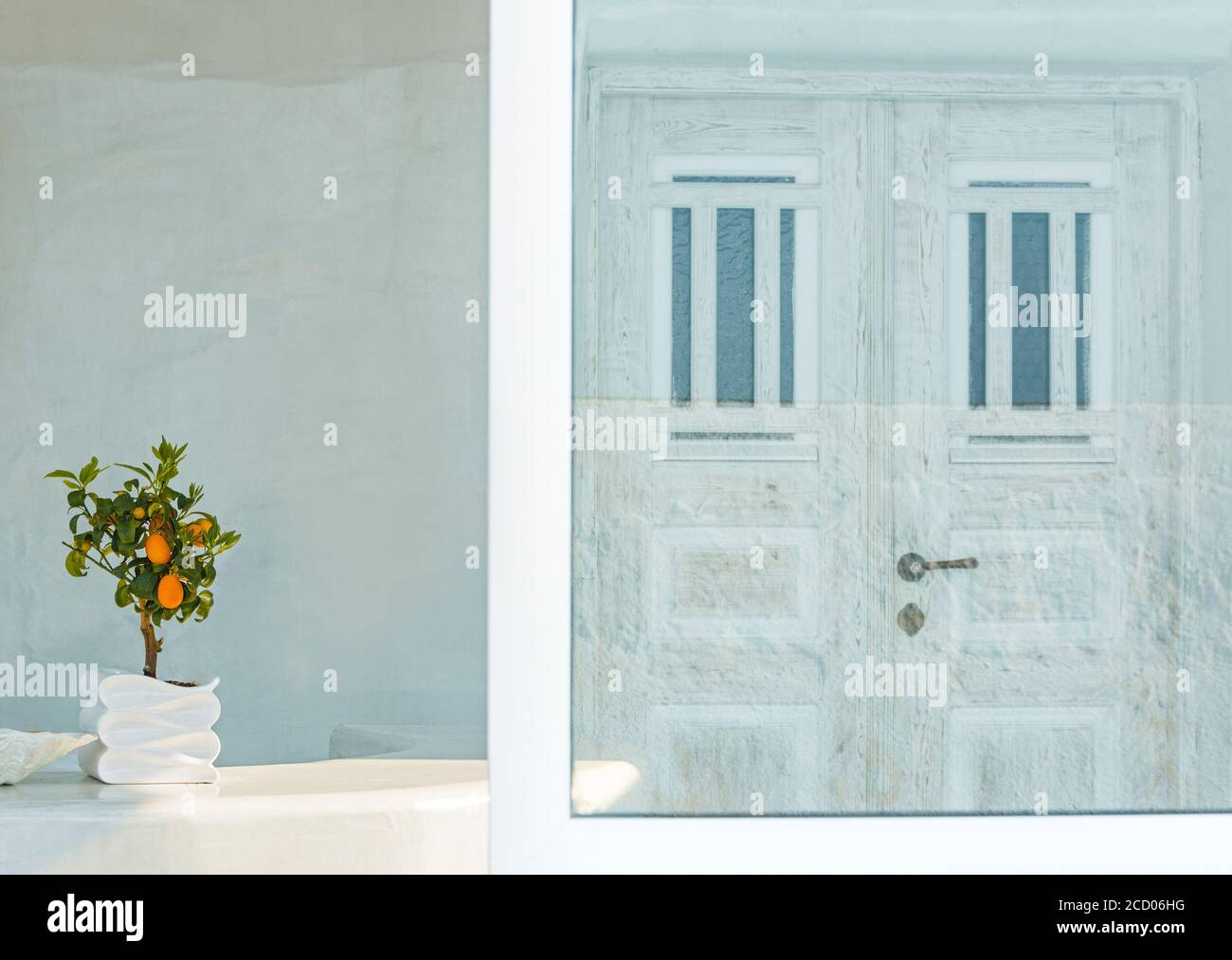 kykladische Atmosphäre in der Stadt Oia, Insel Santorini, mit einem Bonsai-Baum und einer weißen Tür, die sich in einem Spiegel entlang des Weges spiegelt Stockfoto