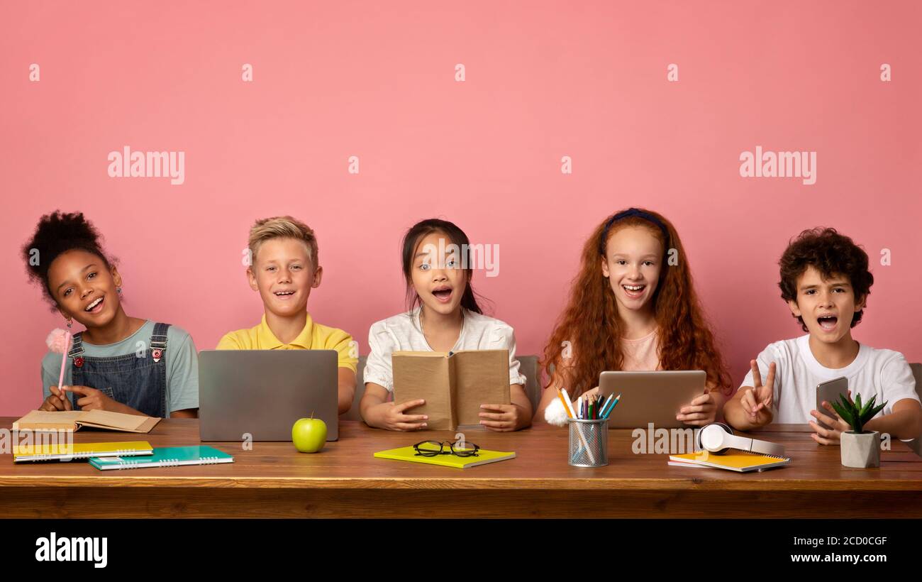 Gruppe von begeisterten Schülern mit Lehrbüchern und Gadgets sitzen am Tisch und Blick auf die Kamera, rosa Hintergrund. Leerzeichen Stockfoto