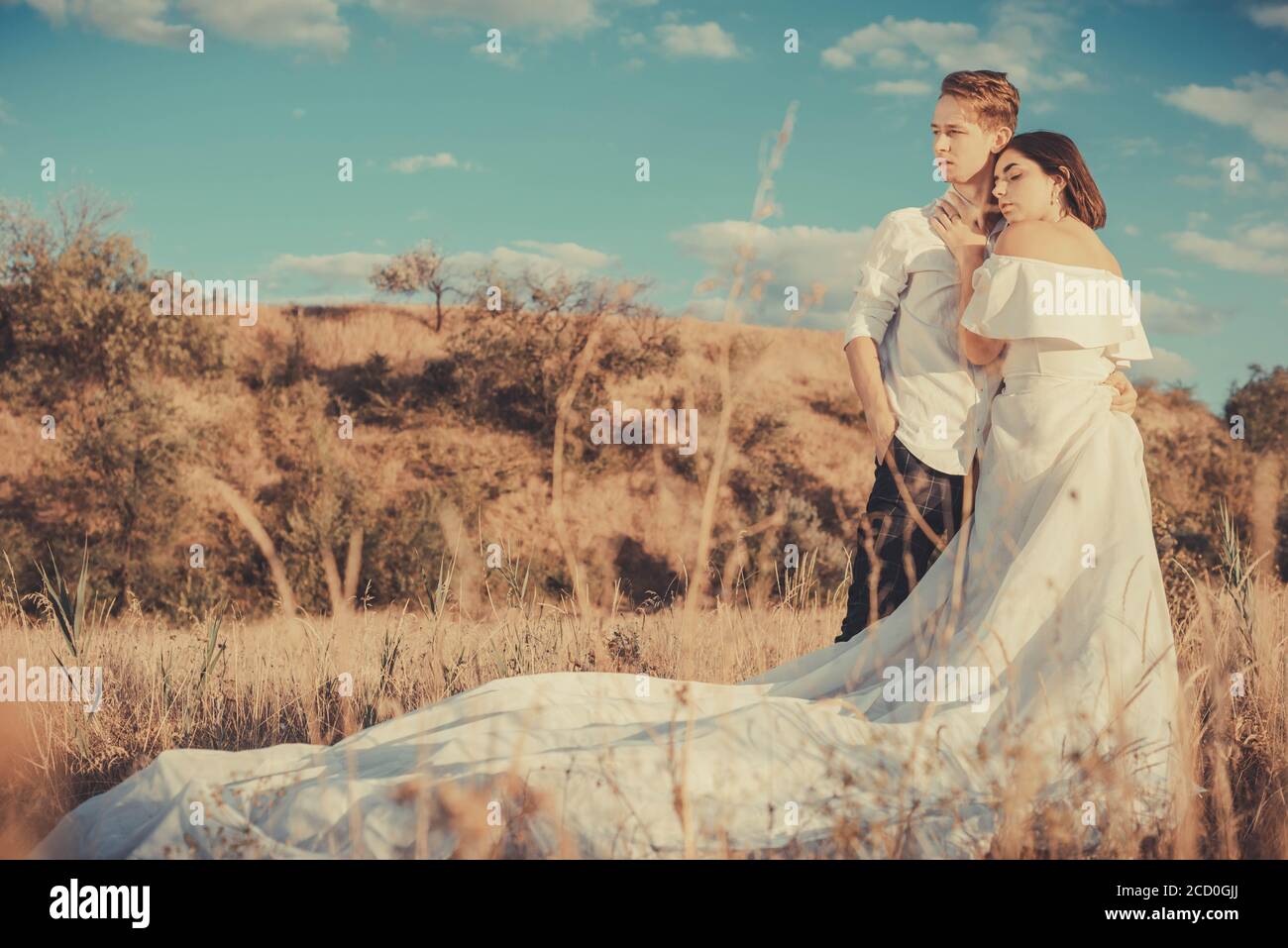 Junge Hochzeit Paar von einem Mann und einem Mädchen, in einem weißen Kleid, auf einem Feld, vor dem Hintergrund eines bewölkten blauen Himmel, Sonnenuntergang. Liebe zum jungen Peop Stockfoto