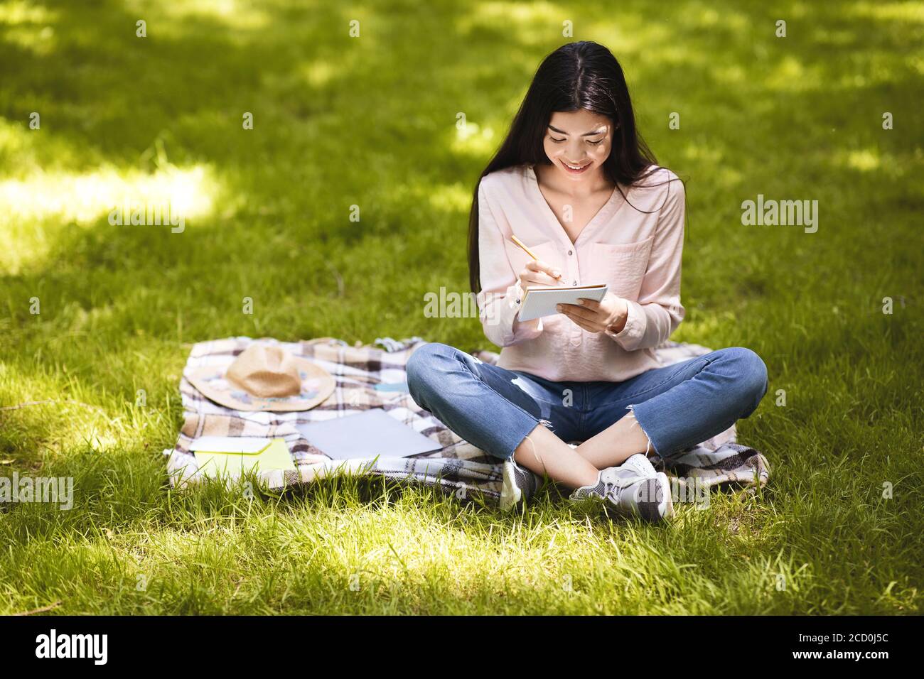 Lächelndes asiatisches Mädchen, das Notizen in Notizblock macht, während es draußen sitzt Im Park Stockfoto