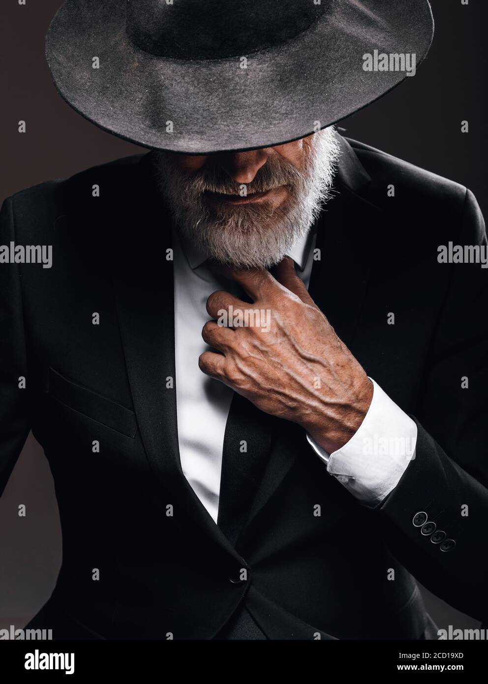 Ein alter männlicher Schauspieler, der eine englische Spionagerolle spielt, in schwarzem Anzug gekleidet, versteckt hinter Fedora-Hut, mit geheimnisvollem eleganten Look. Isoliertes Studio portra Stockfoto