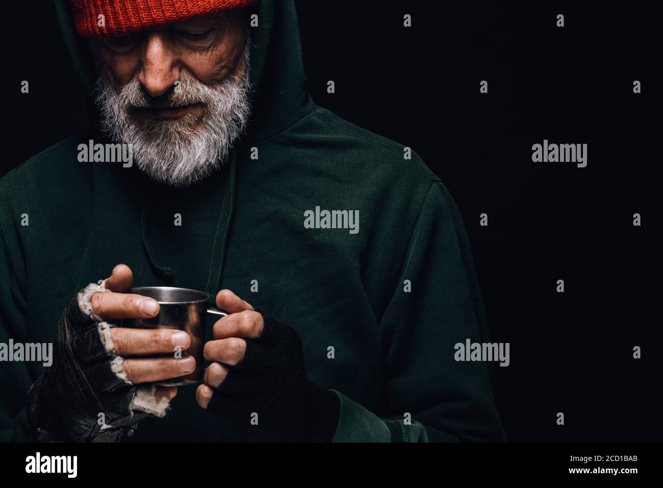 Alter obdachloser Mann mit grauem Bart, der sich grün bedeckt Dekrepit tragen eine Tasse heißen Tee zu warm Sich selbst in einer kalten Nacht Stockfoto