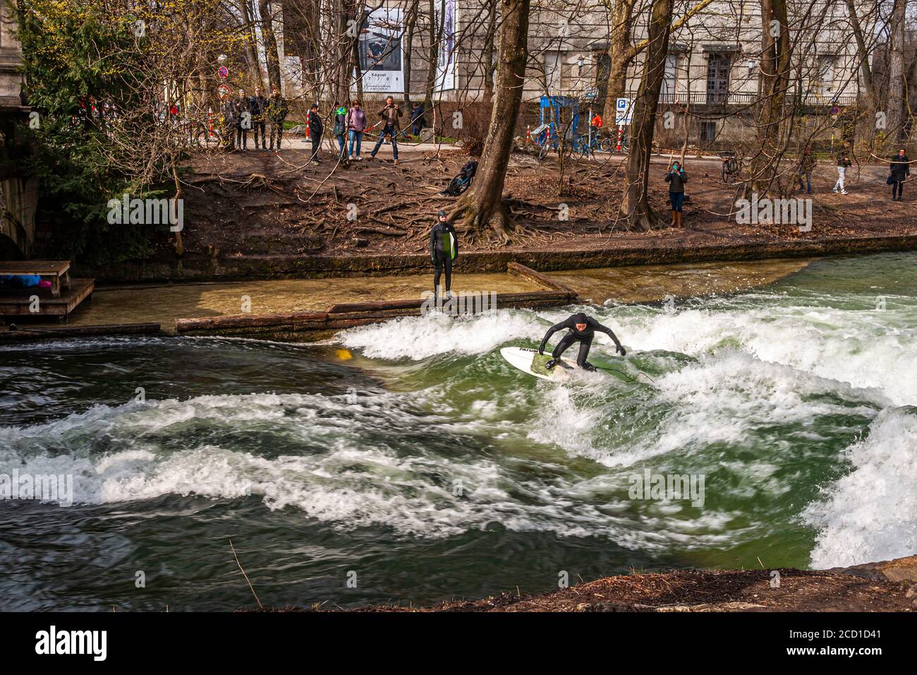 River-Surfing auf dem Eisbach in München, Deutschland. Die stehende Welle kann so lange gesurft werden, wie das Gleichgewicht hält, und in geschäftigen Zeiten bildet sich eine Schlange von Surfern am Ufer Stockfoto