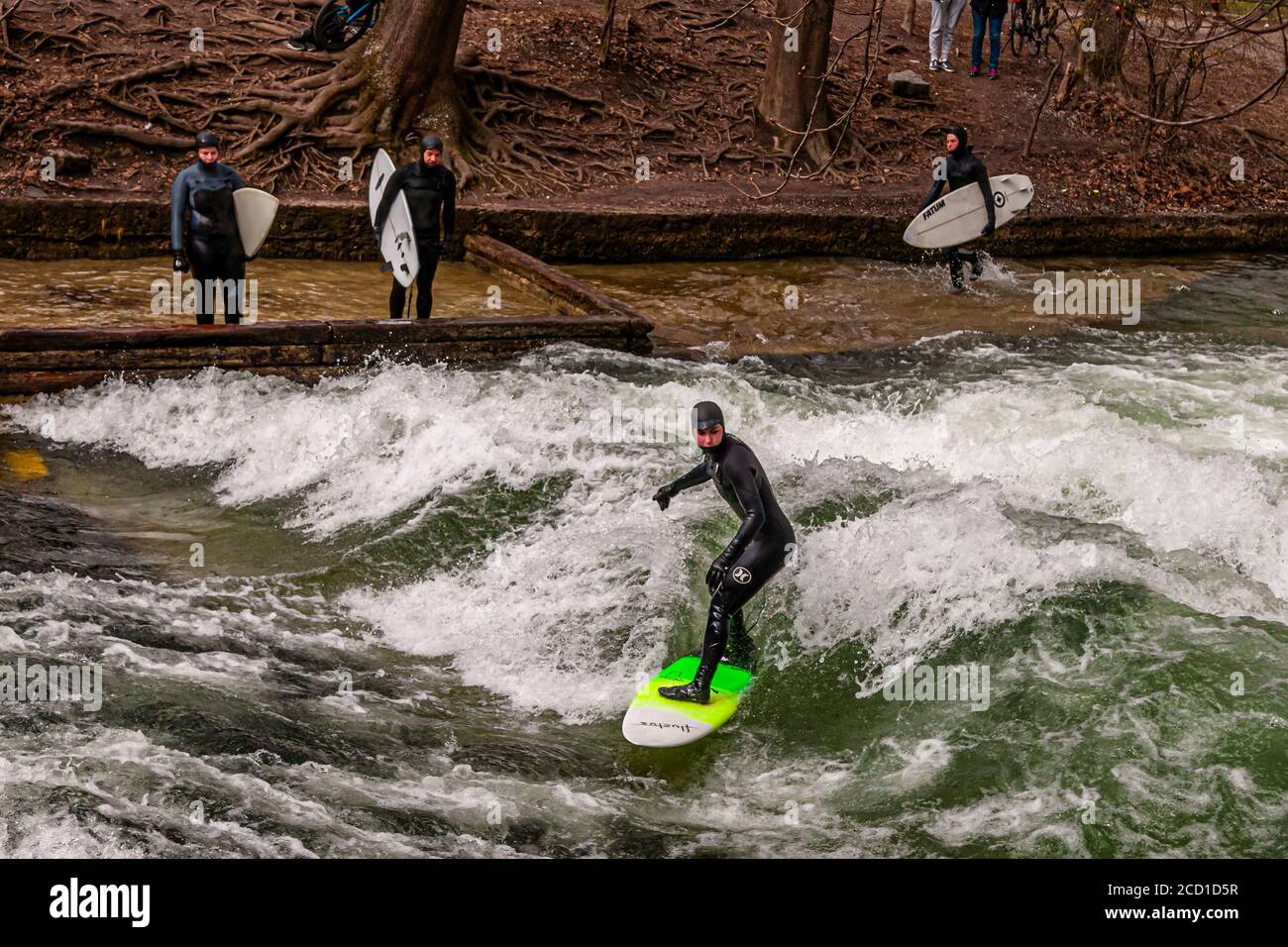 River-Surfing auf dem Eisbach in München, Deutschland. Die stehende Welle kann so lange gesurft werden, wie das Gleichgewicht hält, und in geschäftigen Zeiten bildet sich eine Schlange von Surfern am Ufer Stockfoto