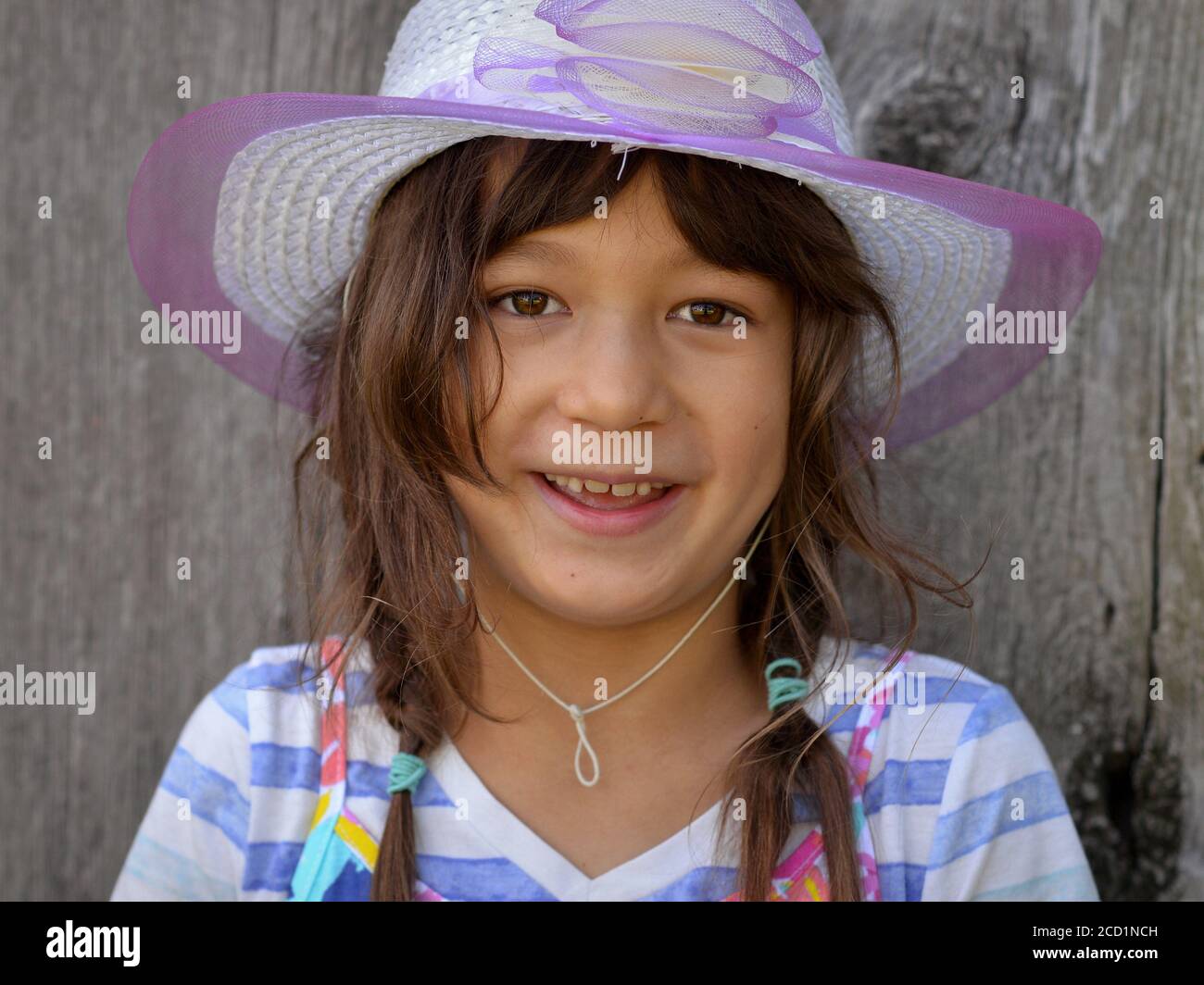 Niedliches kleines Mixed-Race-Mädchen (kaukasisch und südostasiatisch) mit langen, ungepflegten Haaren trägt einen modernen Sonnenhut und lächelt in die Kamera. Stockfoto
