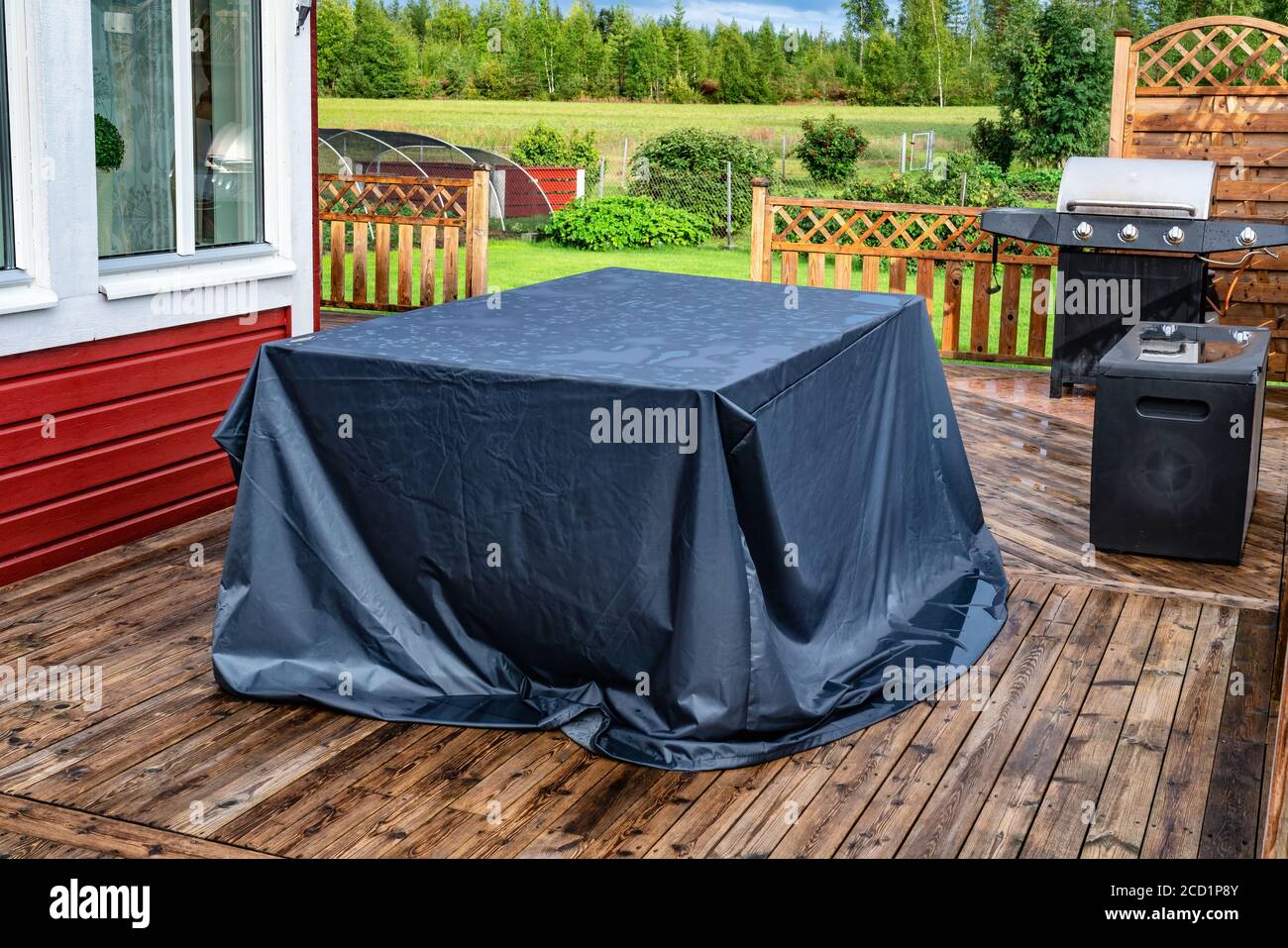 Auf der Holzterrasse steht ein großer, gegen Regen verdeckter Tisch. Rotes Haus, grüner Garten, Feld und Wald im Hintergrund. Nasse Terrasse nach starkem Regen Stockfoto