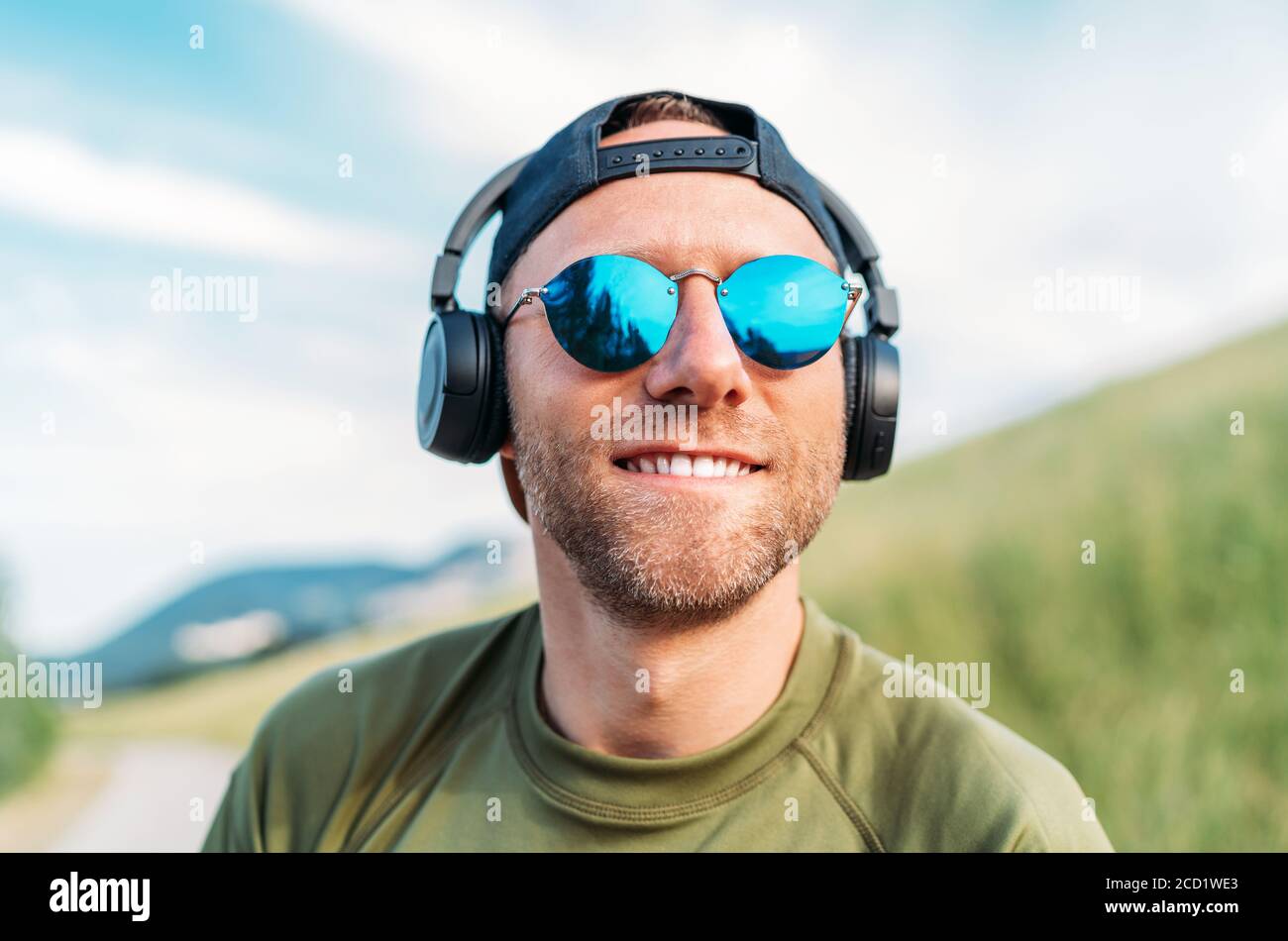 Cooler Mann in einer Baseballmütze, kabellosen Kopfhörern und blauer Sonnenbrille lächelt und genießt Lieblingsmusik beim Spielen. Stockfoto