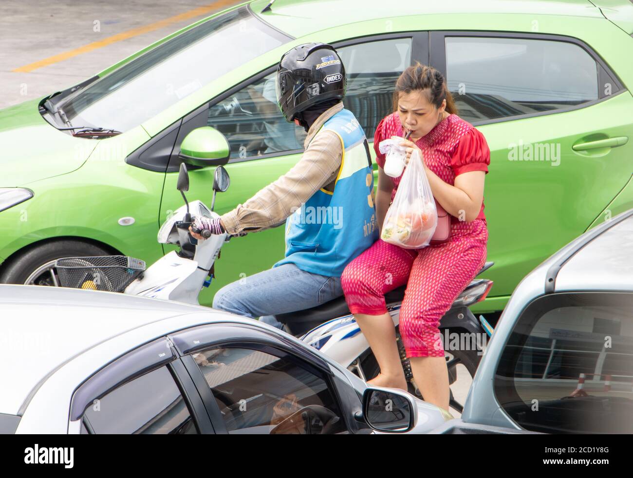 SAMUT PRAKAN, THAILAND, JULI 03 2020, EIN Taxifahrer auf einem Motorrad fährt mit einer trinkenden Frau. Der mototaxi-Fahrer trägt einen Passagier zwischen den Autos auf Stockfoto
