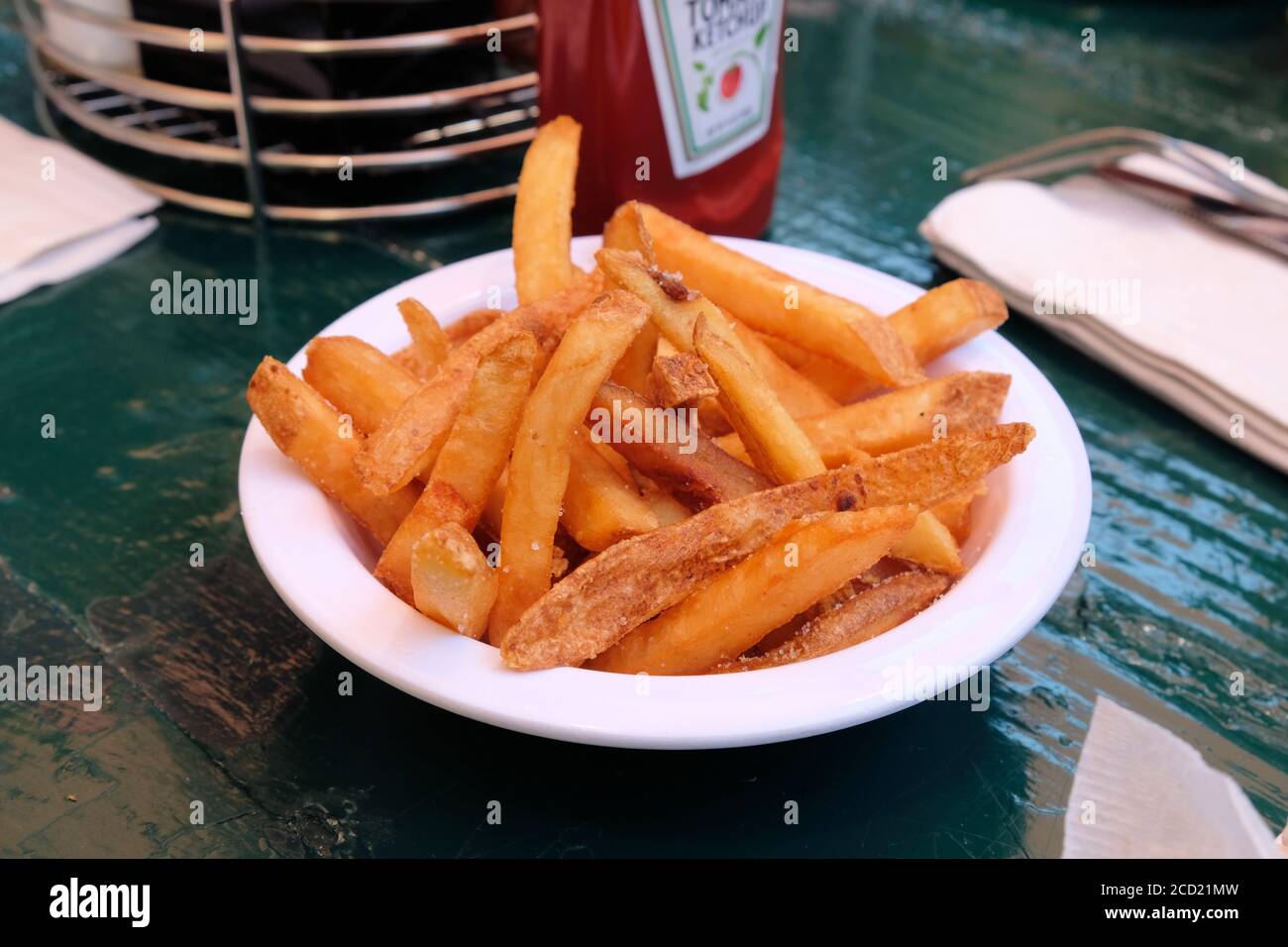 Nahaufnahme einer amall weißen Schüssel mit Steak Pommes oder Pommes auf einem grünen Picknicktisch und einer verschwommenen Ketchup-Flasche im Hintergrund; Beilage. Stockfoto