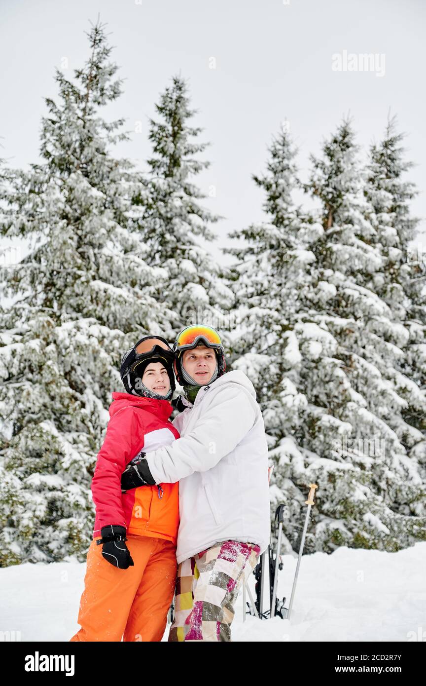 Charmanter Mann und Frau in Skianzügen und Helmen umarmen und beiseite schauen, während im Winterwald mit schneebedeckten Nadelbäumen auf dem Hintergrund stehen. Mann Skifahrer umarmt schöne junge Frau und lächelnd. Stockfoto