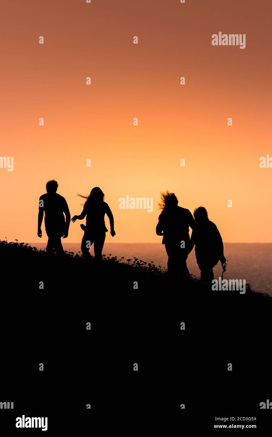 Vier Menschen wandern auf dem Pentire Point East, der von einem intensiven, farbenfrohen Sonnenuntergang in Newquay in Cornwall umgeben ist. Stockfoto