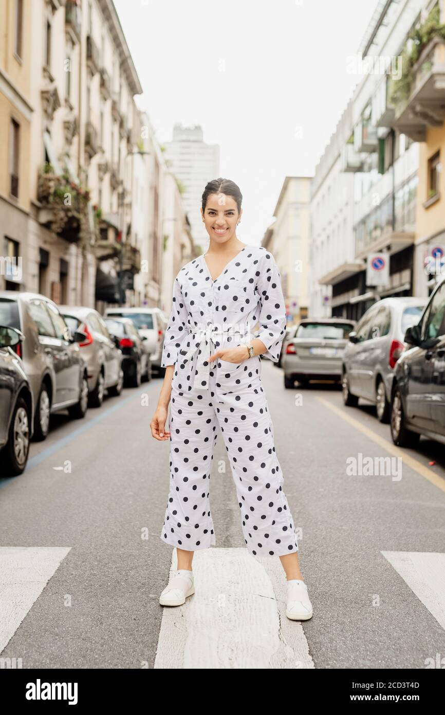 Portrait einer lächelnden Frau in weißem und schwarzem Tupfenjumpsuit, auf Fußgängerüberweg stehend. Stockfoto