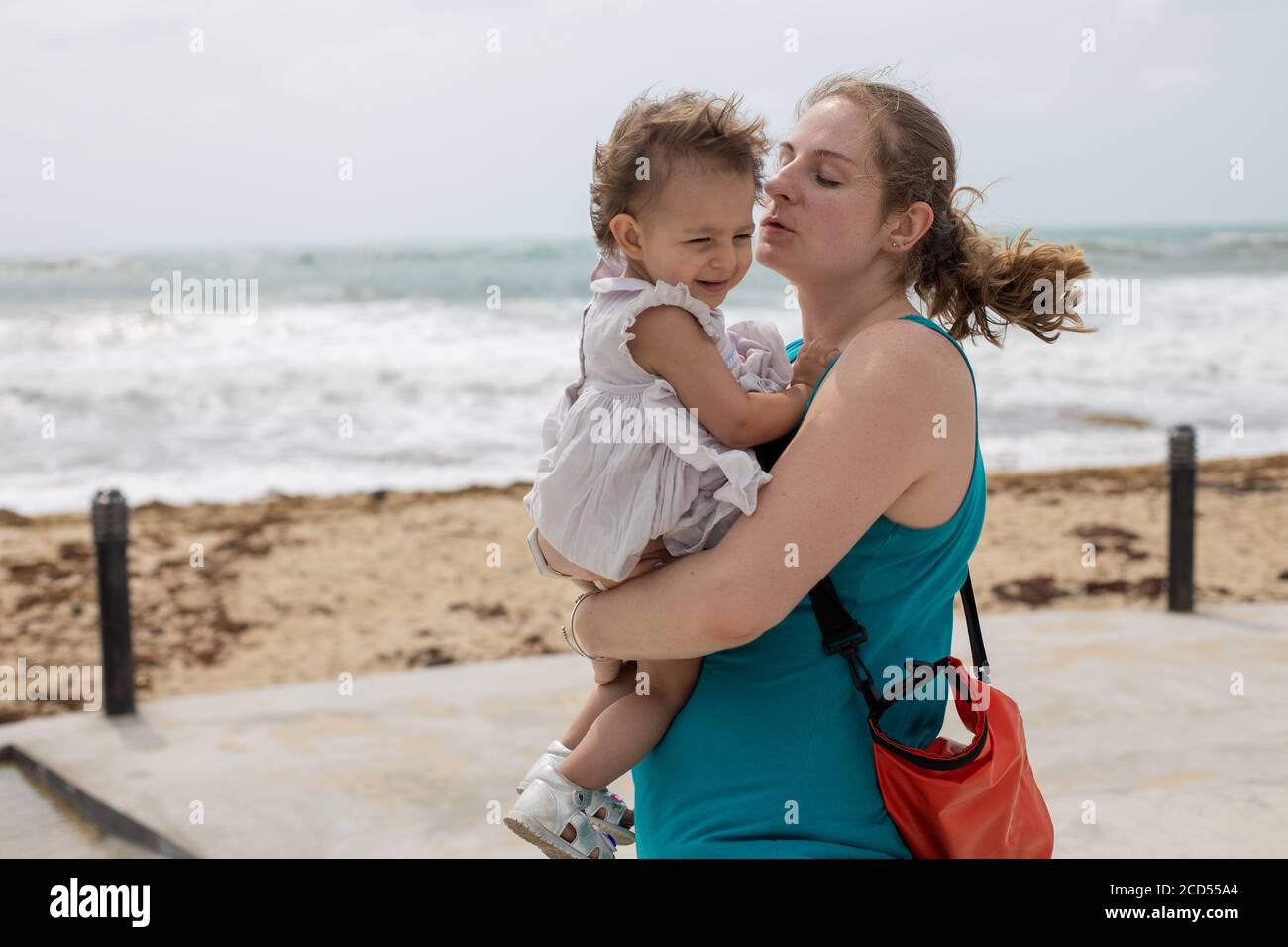 Eine junge Mutter hält ein kleines Mädchen in den Armen vor dem Hintergrund des Meeres in einem Sturm, starken Wind Stockfoto