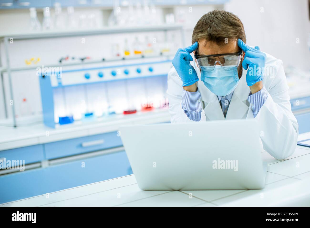 Hübscher junger Forscher in weißem Labormantel und Schutzmaske Arbeiten mit einem Laptop im Labor Stockfoto