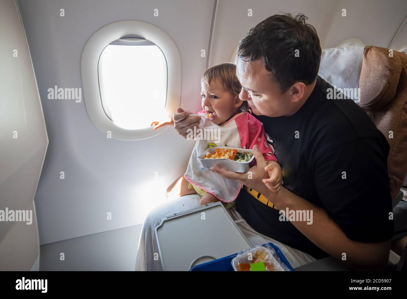 Mahlzeit an Bord der aircraftl Economy Class. Vater füttert das kleine Kleinkind im Flugzeug mit speziellen Mahlzeiten. Stockfoto